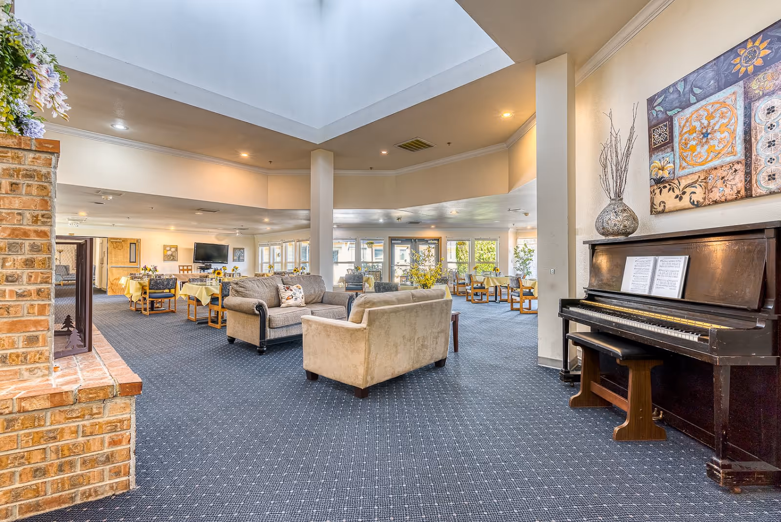 Spacious common area in an assisted living facility featuring a piano with sheet music, a beige armchair and sofa, a brick fireplace, and several dining tables with yellow tablecloths. The room has large windows allowing natural light and a high ceiling with recessed lighting.