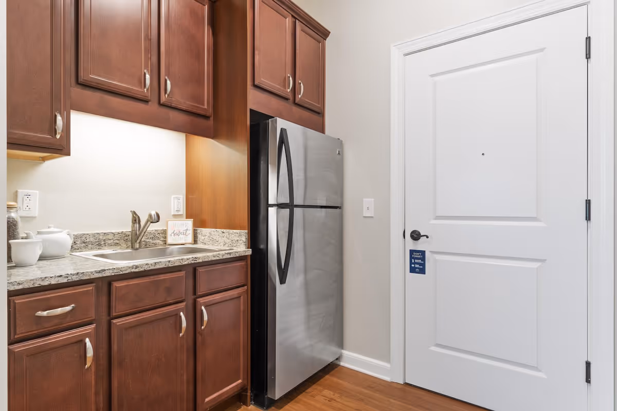 A small kitchen area featuring dark wooden cabinets, a granite countertop with a sink, a stainless steel refrigerator, and a white door with a black handle. There is a small decorative sign on the countertop that reads 'Home Sweet Home'.