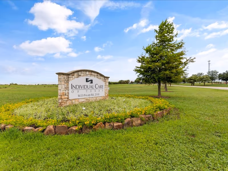 Stone entrance sign for Individual Care of Texas sits in a landscaped grassy median with trees under a blue sky.