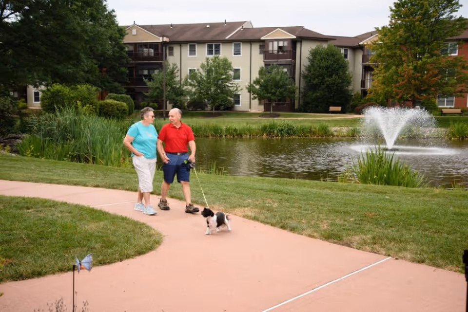 An elderly couple walking a small black and white dog on a leash along a paved path beside a pond with a water fountain. Behind them are trees, bushes, and a multi-story residential building.