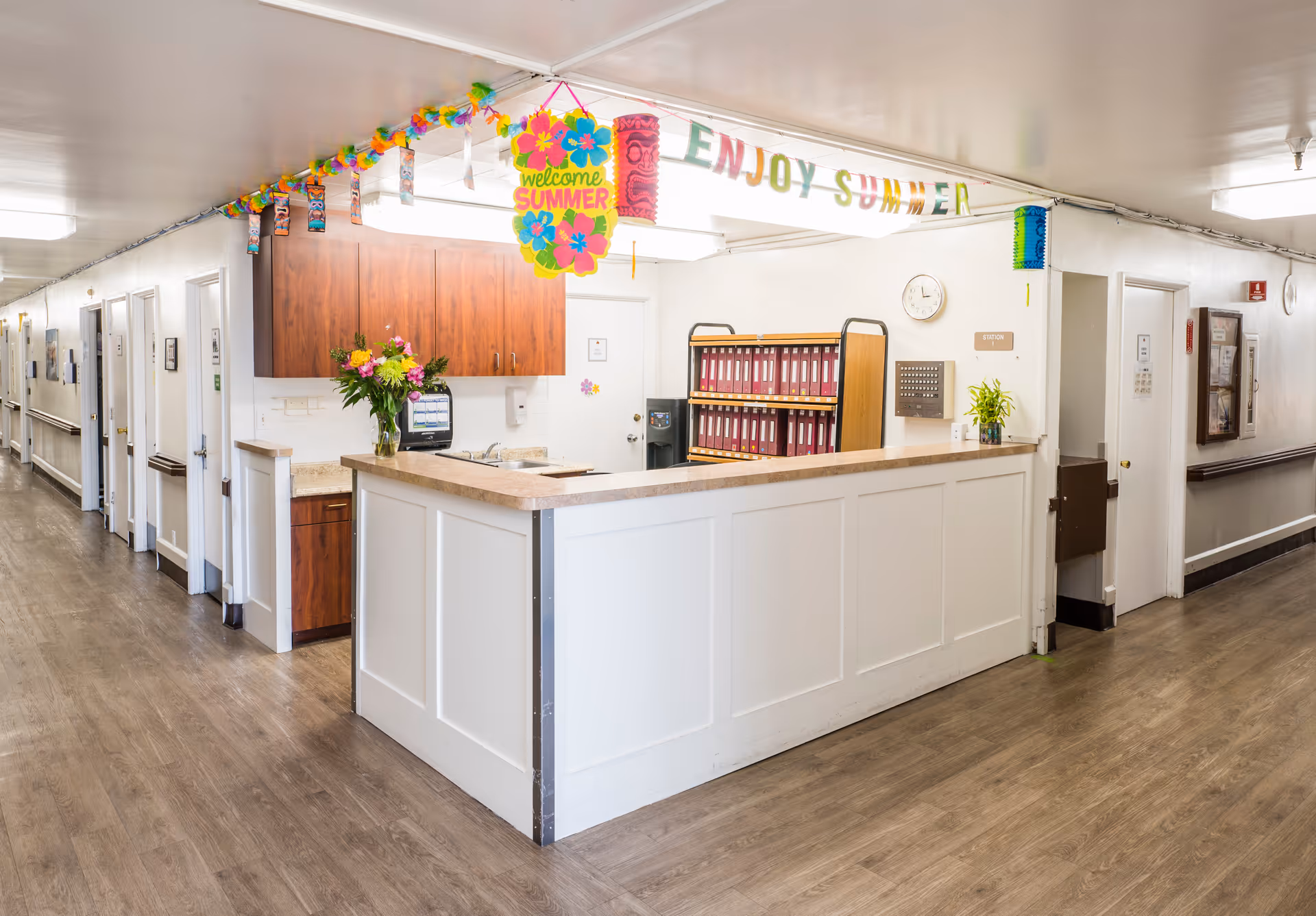 A bright hallway in a senior living facility with a reception desk in the center. The desk has a vase with flowers and a computer monitor. Above the desk, colorful summer-themed decorations hang from the ceiling, including a sign that says 'welcome SUMMER' and another that says 'ENJOY SUMMER'. The hallway has multiple doors along the walls and wood-style flooring.