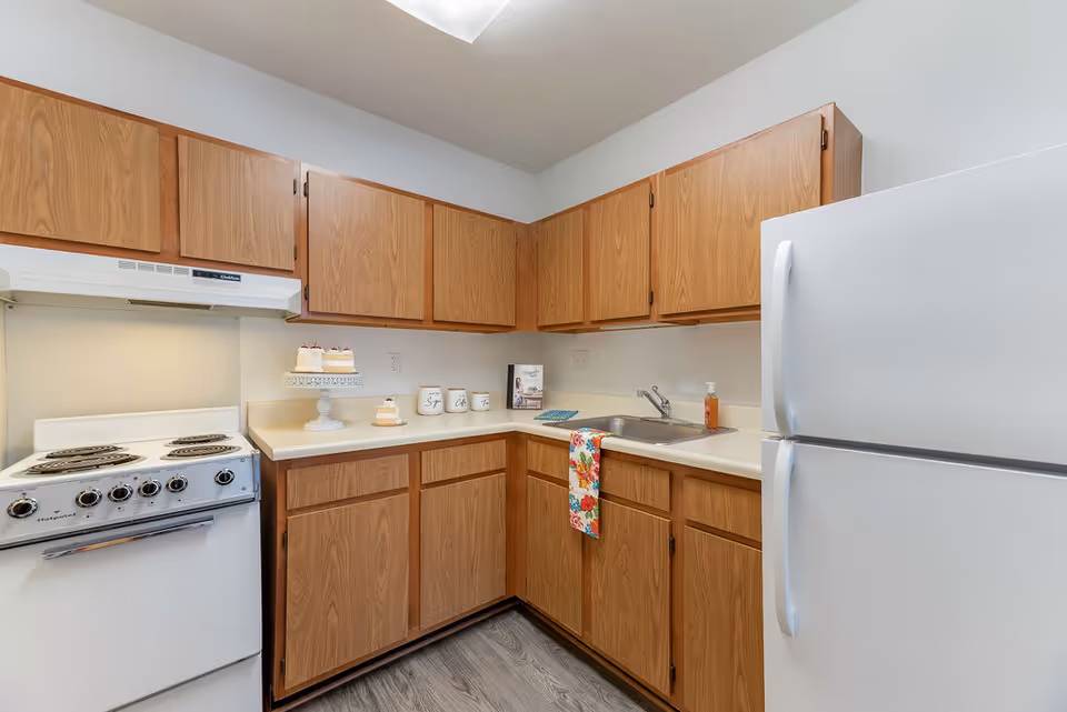 A small kitchen with wooden cabinets, a white electric stove with four burners, a white refrigerator, a stainless steel sink with a colorful floral towel hanging from the counter, and a few decorative items including a cake stand with cupcakes and three labeled canisters on the countertop.