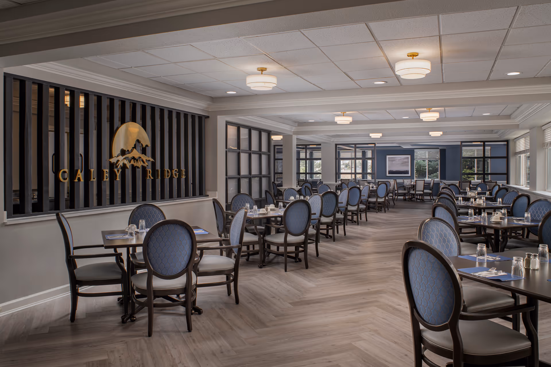 A spacious and well-lit dining room at Caley Ridge Assisted Living with multiple tables and chairs arranged neatly. The chairs have blue cushioned backs and seats, and the tables are set with glasses, napkins, and silverware. The room features large windows allowing natural light, modern ceiling lights, and a decorative wall with the Caley Ridge logo.