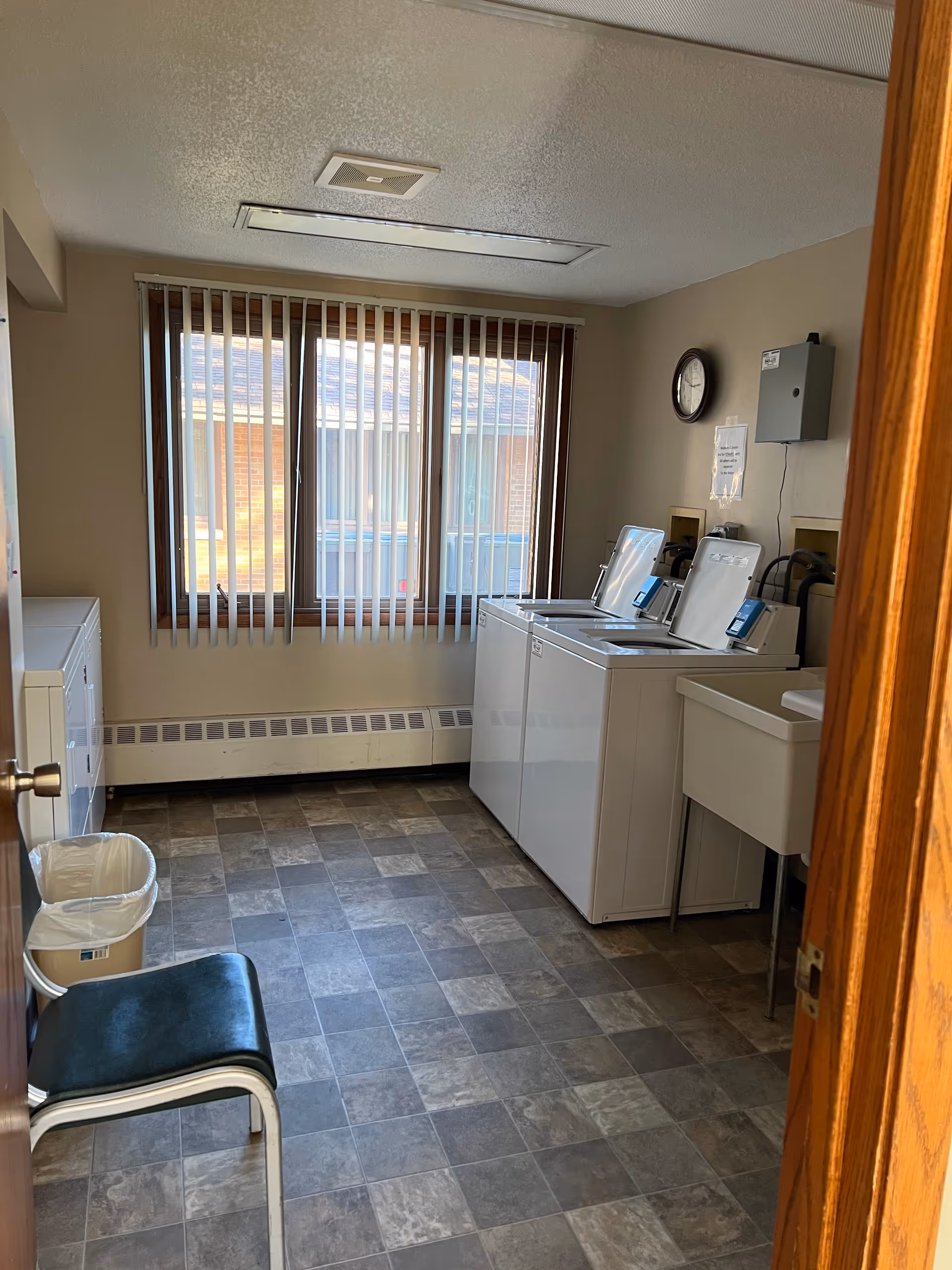 A laundry room with two top-loading washing machines, a utility sink, a chair, a trash can, and a window with vertical blinds. The floor has a checkered tile pattern, and there is a clock and a notice on the wall above the machines.