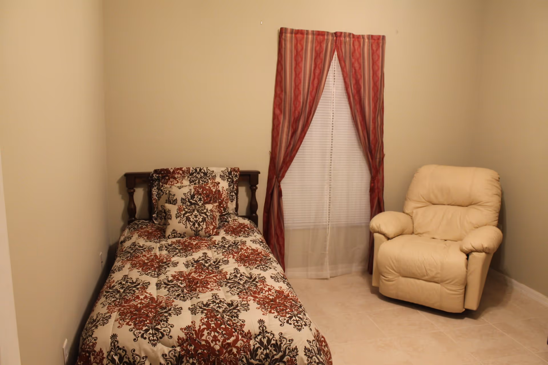 A small bedroom with a single bed covered in a patterned comforter and pillows in shades of red, black, and white. Next to the bed is a beige recliner chair. A window with white blinds and red patterned curtains is centered on the wall behind the chair.