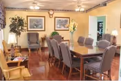 A well-lit dining room with a long wooden table surrounded by eight striped upholstered chairs. The room features hardwood floors, two framed landscape paintings on the wall, a potted plant in the corner, and a vase with flowers on the table. There are ceiling fans with lights and a doorway leading to another room.