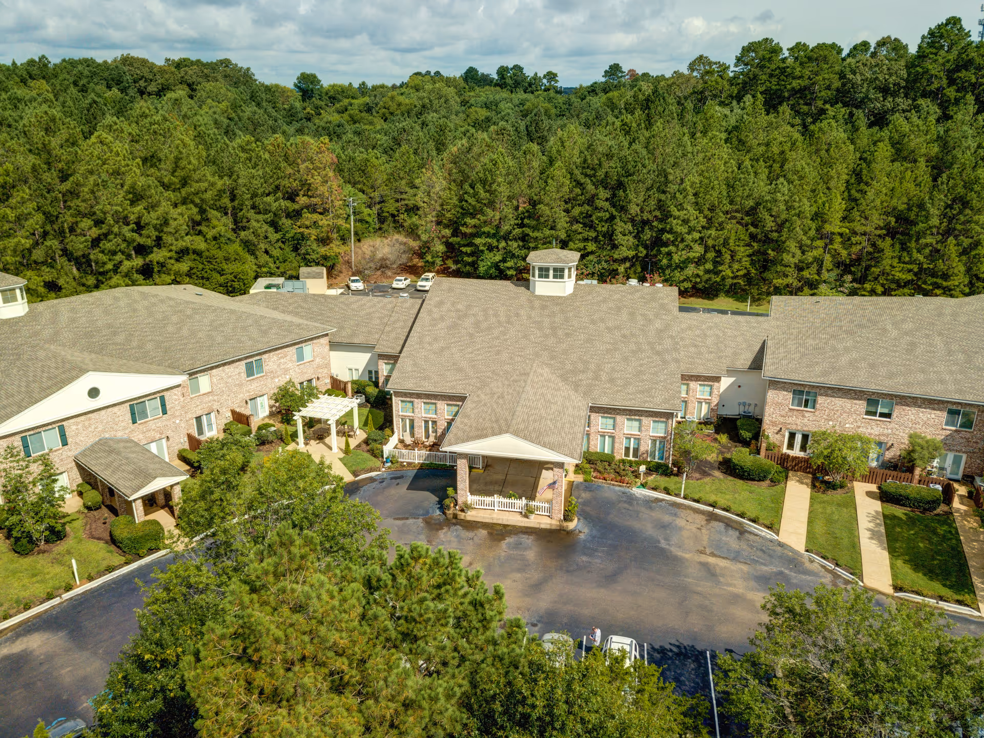Aerial view of Castlewoods Place senior living facility surrounded by dense green trees. The building has a large covered entrance with a circular driveway and several parked cars. The facility is made of brick with multiple windows and landscaped greenery around the pathways.