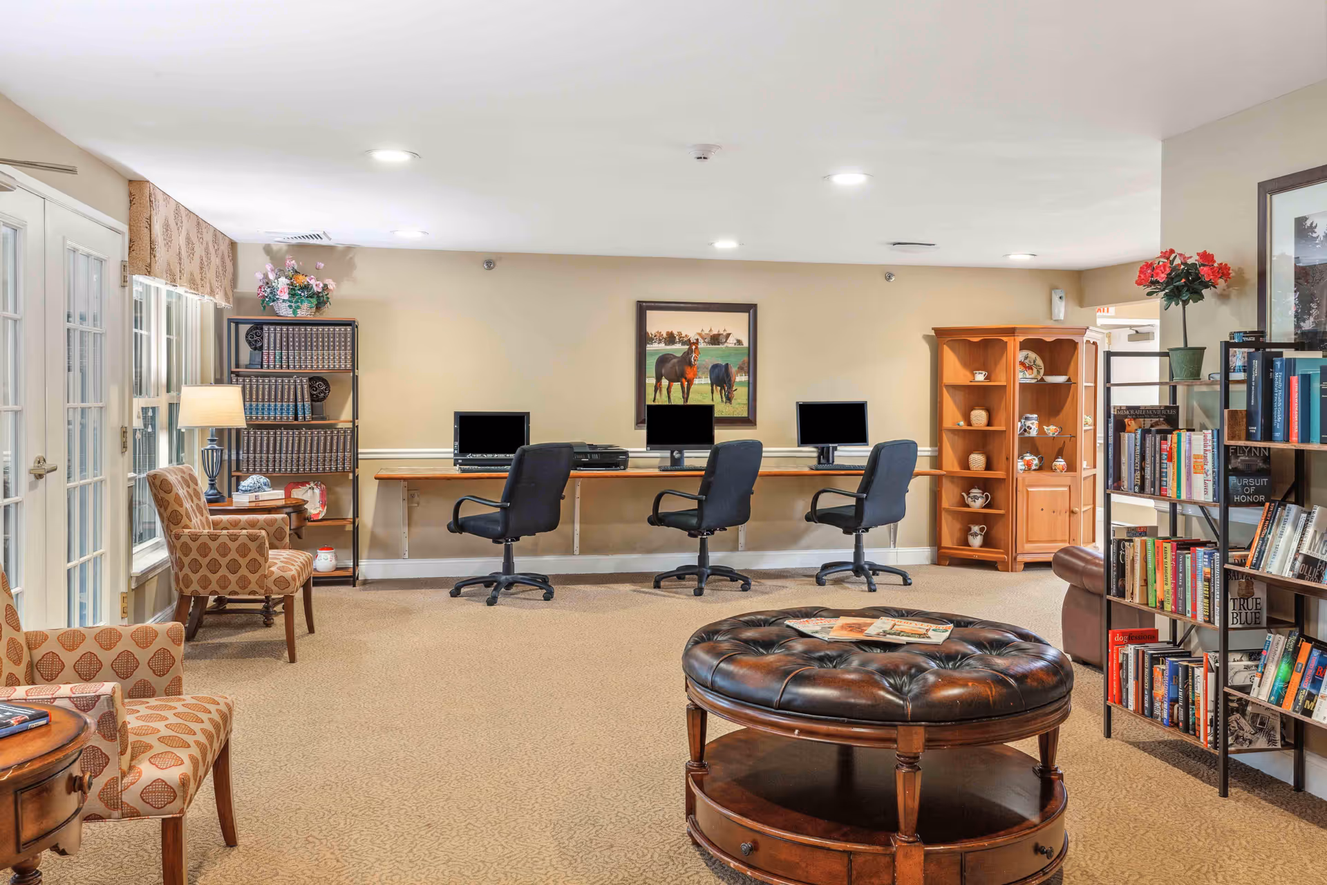 A cozy senior living common area with three computer workstations along the back wall, each with a black office chair. The room features beige walls and carpet, a round tufted leather ottoman in the center, bookshelves filled with books and decorative items, and comfortable patterned armchairs near a window with French doors. A framed painting of horses hangs on the wall above the computers.