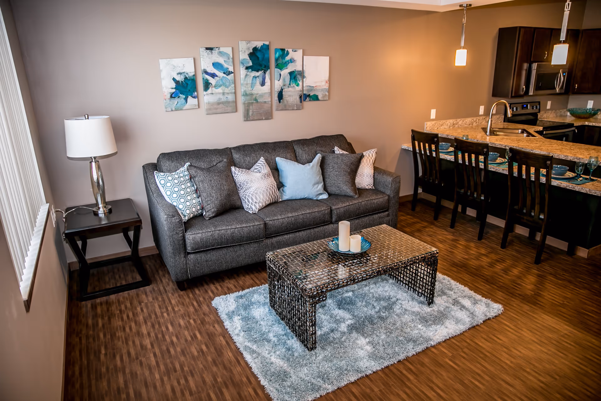 A cozy living room area with a gray sofa adorned with multiple patterned cushions, a glass-top coffee table on a light blue shag rug, a side table with a lamp, and a kitchen counter with four chairs and place settings in the background.