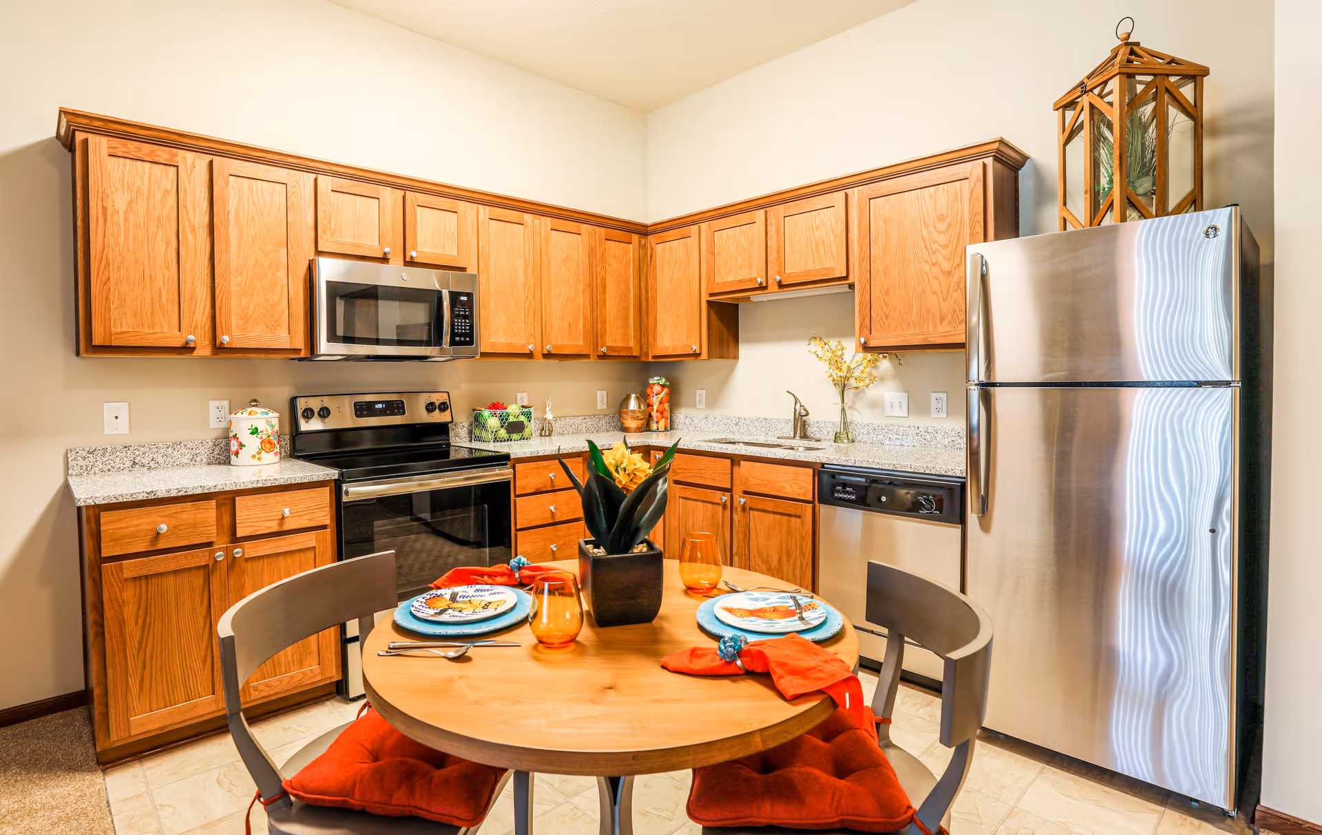 A kitchen with wooden cabinets, stainless steel appliances including a refrigerator, microwave, stove, and dishwasher. A round wooden dining table with two chairs, each with a red cushion, is set with plates, orange glasses, and red napkins. A decorative plant is placed in the center of the table.