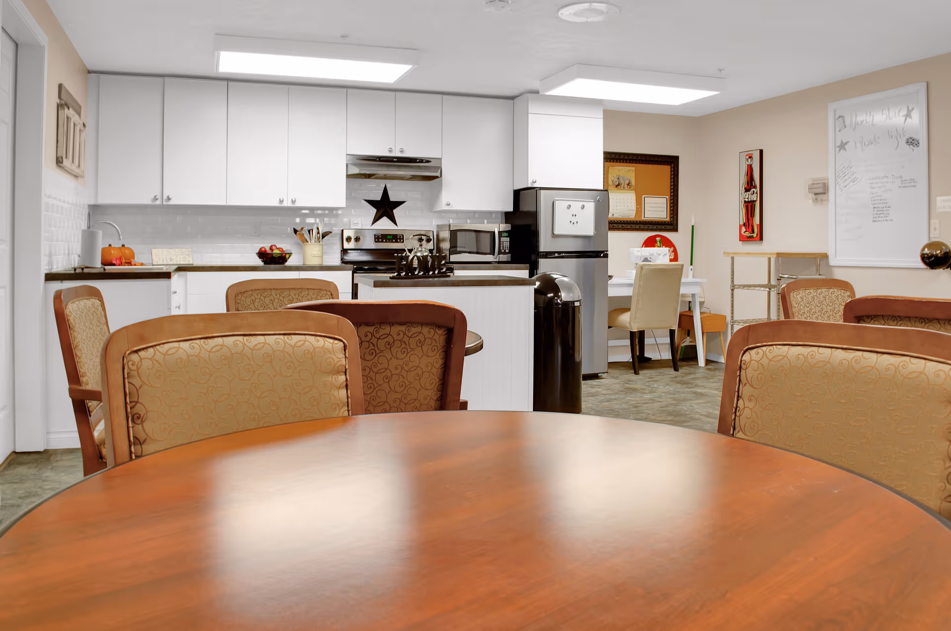 Communal dining area with round wooden tables and upholstered chairs facing a white kitchenette in a senior living facility.