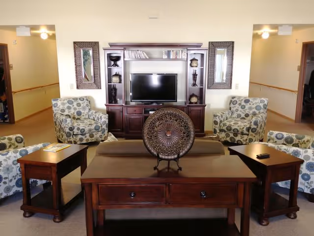A cozy living room area in an assisted living facility featuring a dark wooden entertainment center with a flat-screen TV, two decorative mirrors on either side, and shelves with decorative items. The room has four patterned armchairs arranged around a wooden coffee table with a decorative circular metal piece on it, and two matching wooden side tables.
