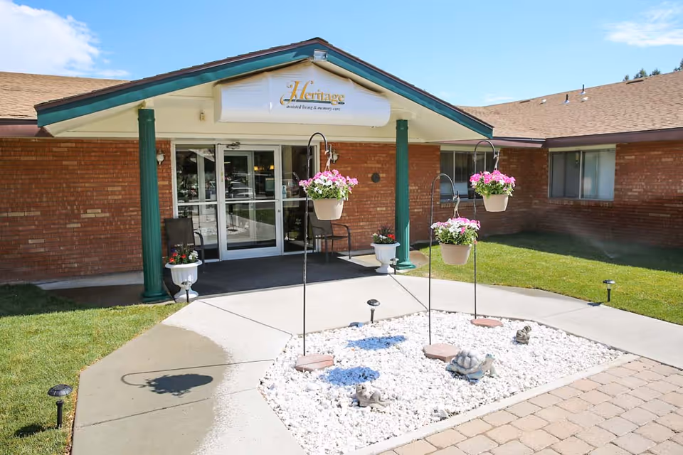 Entrance of Heritage Assisted Living Of Twin Falls facility with a covered porch supported by green columns, glass double doors, hanging flower pots with pink and white flowers, and a landscaped area with white rocks and small garden statues.