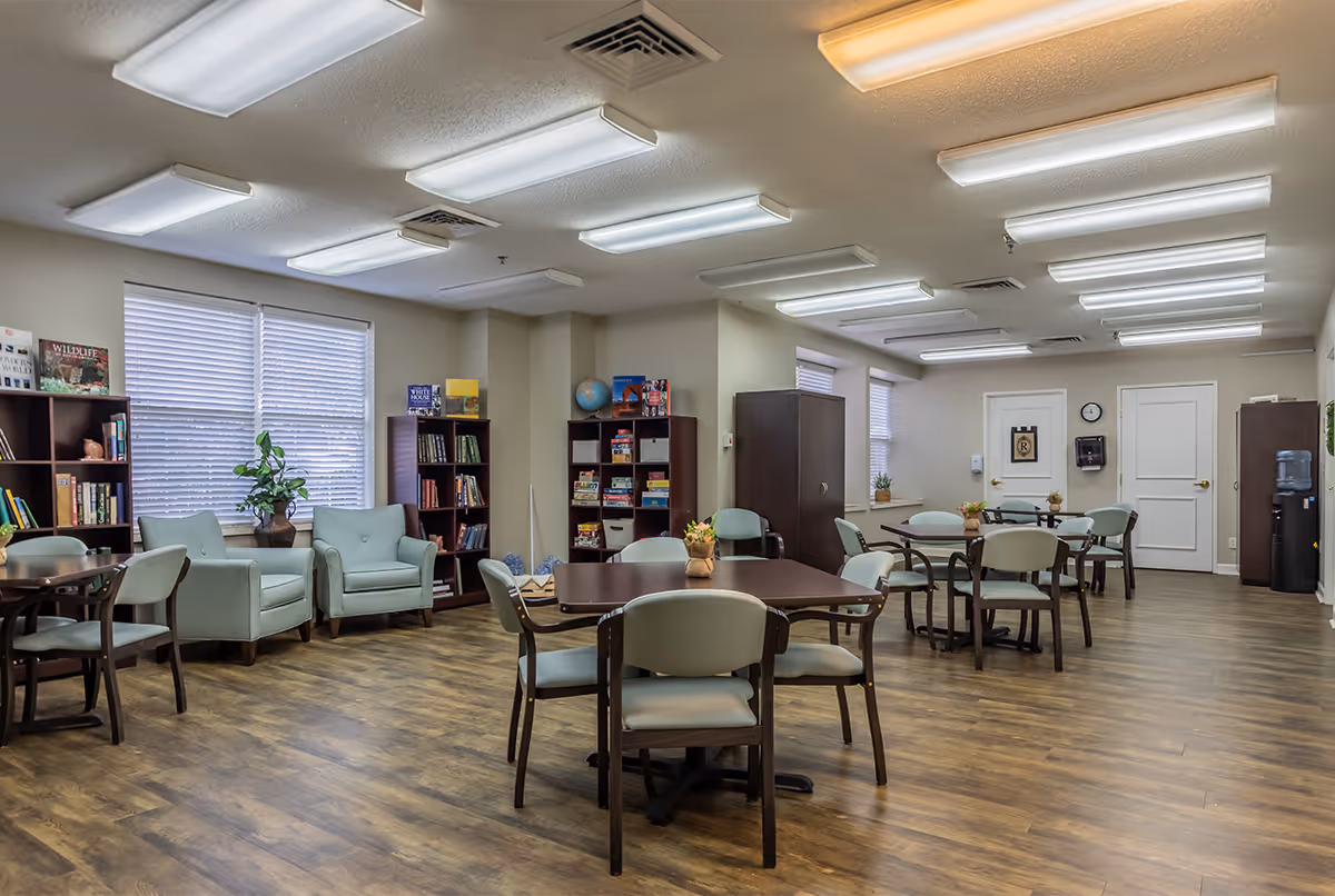 A well-lit common area with multiple tables and chairs arranged for group seating. The room features wooden flooring, bookshelves filled with books and games, two armchairs near a window with blinds, and a water cooler in the corner. The walls are light-colored, and the ceiling has multiple fluorescent lights.
