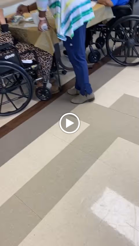 Partial view of a group of people in wheelchairs sitting around a table covered with a yellow tablecloth, with a person standing nearby wearing blue pants and a striped top. The floor has a checkered pattern of beige and brown tiles.