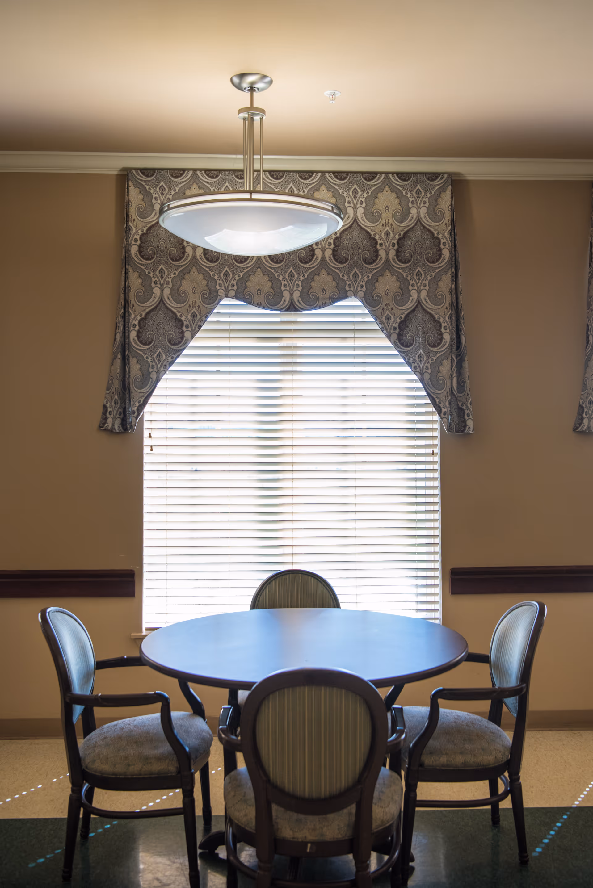 A round wooden table with four cushioned chairs arranged around it in front of a window with closed blinds and decorative valance. A modern ceiling light fixture hangs above the table.