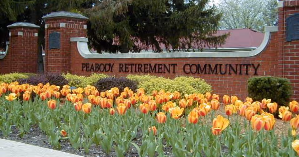 A brick entrance sign for Peabody Retirement Community surrounded by a flower bed filled with orange and yellow tulips and some green shrubs, with trees in the background.