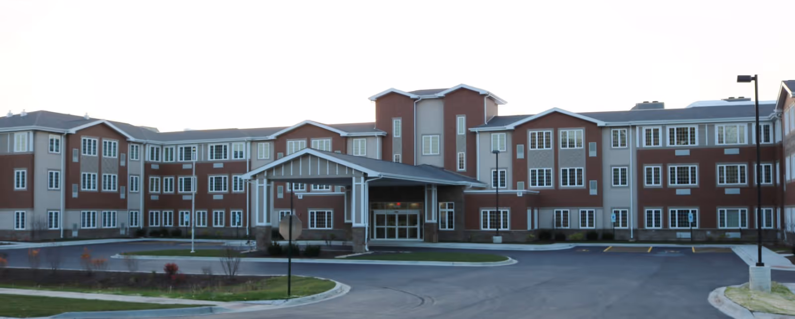 Front exterior view of a large three-story supportive living facility building with multiple windows, a covered entrance, and a circular driveway in front. The building is surrounded by a paved parking area and some landscaped greenery.
