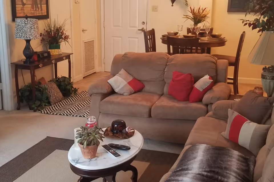 A cozy living room area with two beige sofas adorned with red and beige pillows. A small oval marble-top coffee table in front holds a potted plant, a remote control, and decorative items. In the background, there is a wooden dining table set with plates, glasses, and a flower arrangement. The room is warmly lit and decorated with a table lamp, a side table with a lamp and flowers, and wall art.