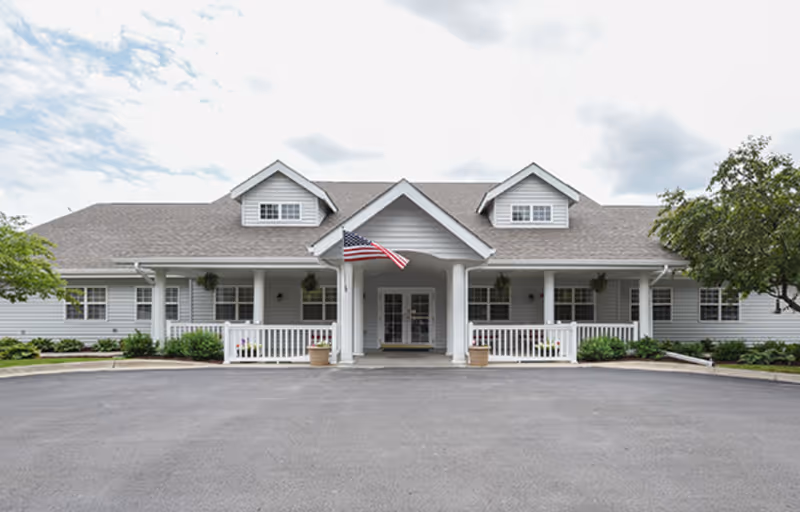 Front exterior view of a single-story senior living facility building with gray siding, a peaked roof, and an American flag hanging above the entrance. There are white railings and potted plants near the entrance, with trees and shrubs on either side.