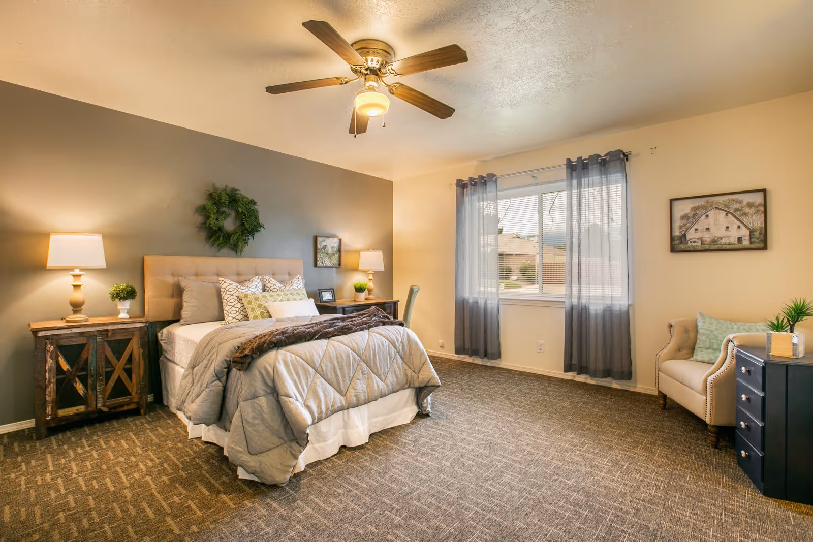 A well-lit bedroom with a large bed featuring multiple pillows and a gray comforter. There are two wooden nightstands with lamps and small plants on either side of the bed. A ceiling fan with a light is mounted above. The room has a large window with sheer gray curtains, a beige armchair with a green pillow, a black dresser with a plant and a tissue box, and framed artwork on the walls. The carpet is patterned in neutral tones.