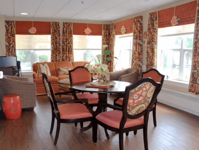 A cozy living room area with a round wooden table surrounded by four upholstered chairs with patterned backs and red seats. Behind the table, there is a comfortable orange sofa and two wicker armchairs. The room has large windows with patterned curtains and red valances, allowing natural light to fill the space. A decorative plant centerpiece is on the table, and the floor is wooden.