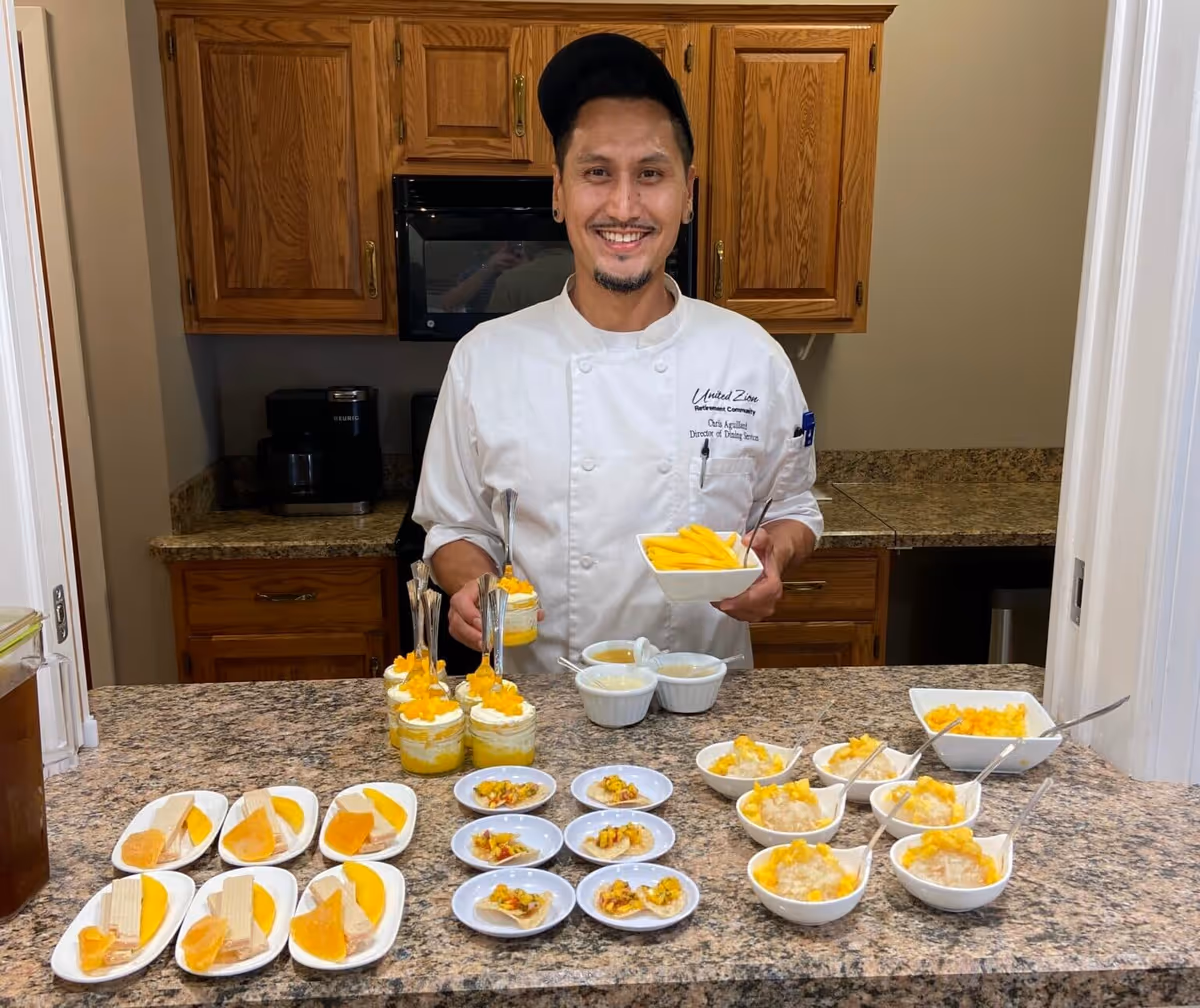 A smiling chef wearing a white uniform and black cap stands behind a kitchen counter with various plated desserts and fruit dishes. The kitchen has wooden cabinets and a granite countertop. The chef is holding a bowl of sliced mangoes.