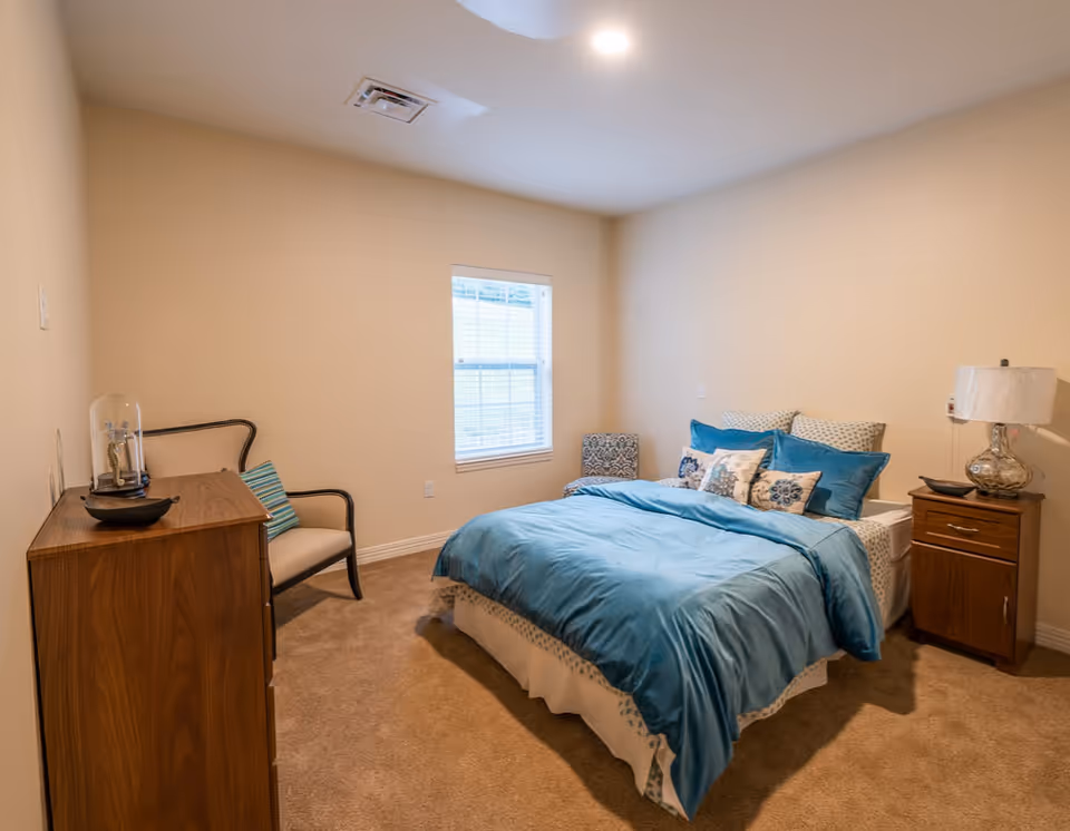 A cozy bedroom with beige walls and carpet, featuring a bed with blue bedding and multiple decorative pillows. There is a wooden nightstand with a glass lamp on the right side of the bed, a wooden dresser on the left side with a decorative item on top, and a cushioned chair with a striped pillow next to the dresser. A window with white blinds is centered on the back wall.