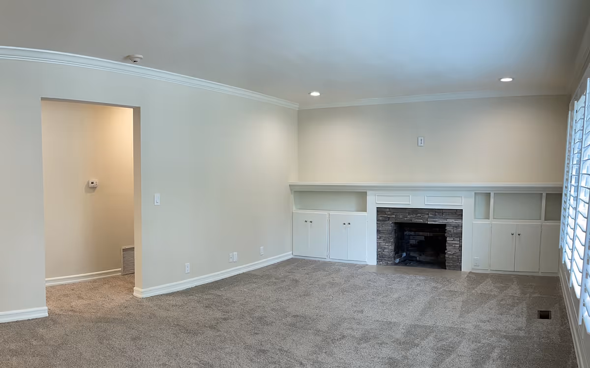 Empty carpeted living room with built-in cabinets and a stone fireplace, recessed lighting, and shuttered windows.