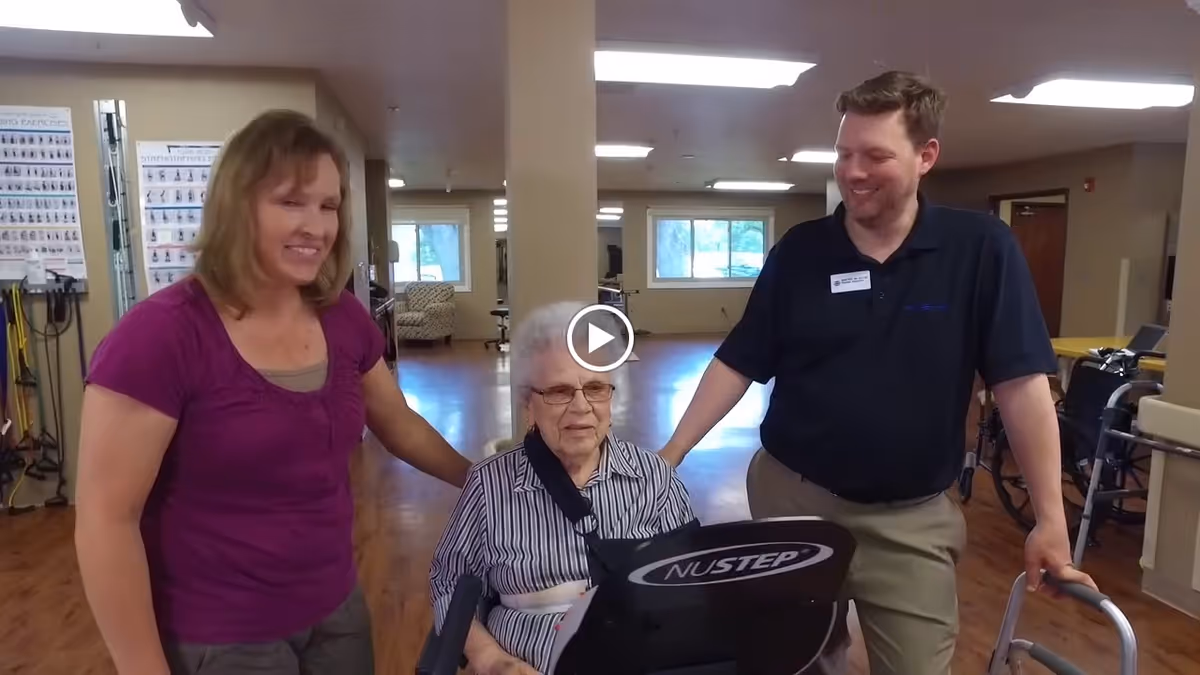 An elderly woman seated on a NuStep exercise machine is assisted by a woman in a purple shirt and a man in a black polo shirt inside a rehabilitation facility with wooden floors and exercise equipment in the background.
