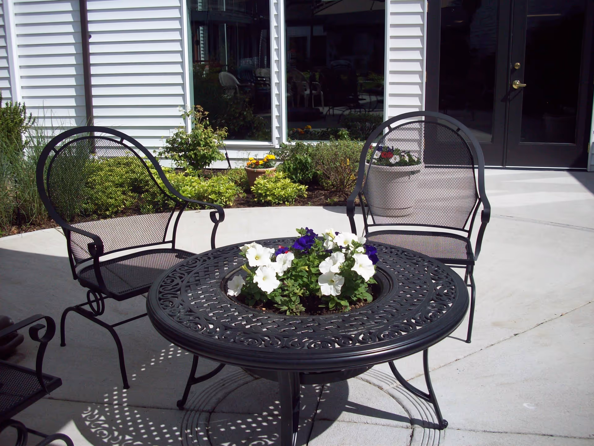 Outdoor patio area with black metal chairs and a round black metal table featuring a built-in planter with white and purple flowers. The patio is adjacent to a building with white siding, windows, and a glass door. There are green shrubs and potted plants near the building.