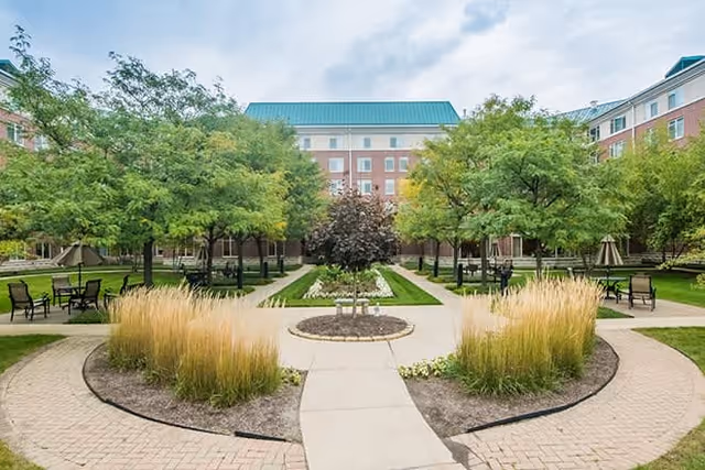 A landscaped outdoor courtyard with a paved walkway leading to a circular garden bed with tall grasses and a small tree in the center. Surrounding the courtyard are green trees, benches, and a multi-story building with a green roof in the background under a cloudy sky.