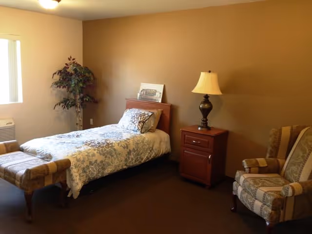 A cozy bedroom with a single bed covered in a floral patterned comforter and pillows. Next to the bed is a wooden nightstand with a decorative lamp. There is a cushioned bench at the foot of the bed and an upholstered armchair with a striped pattern. A potted plant is placed near the window, and the walls are painted in warm beige tones.