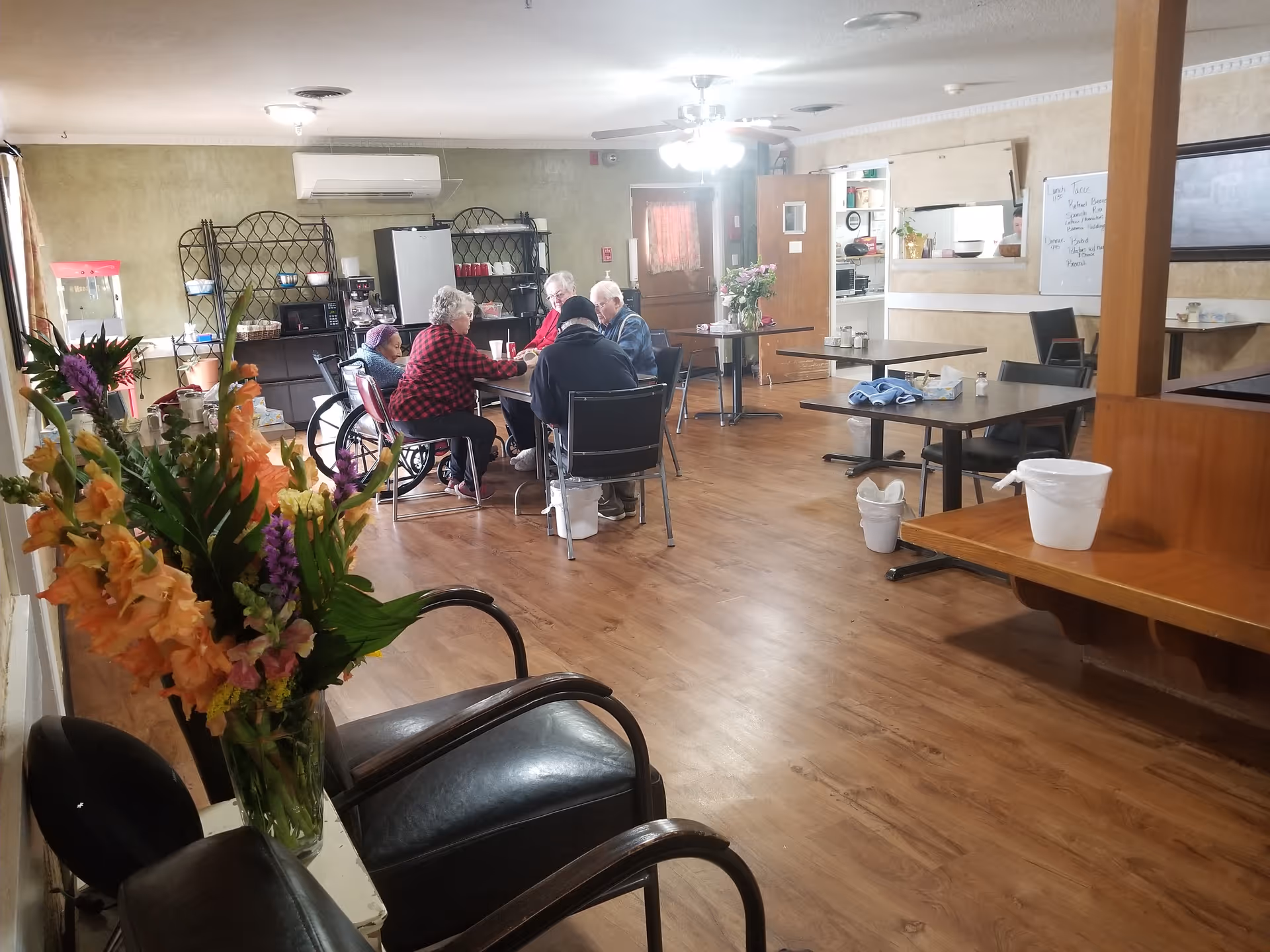 A communal dining/activity room with several residents seated around a table, flowers in the foreground, and empty tables and chairs.