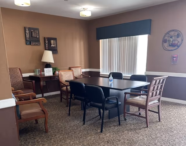 Small communal dining/meeting room with a rectangular table surrounded by various chairs, a lamp on a side table, and wall decor near a window with vertical blinds.