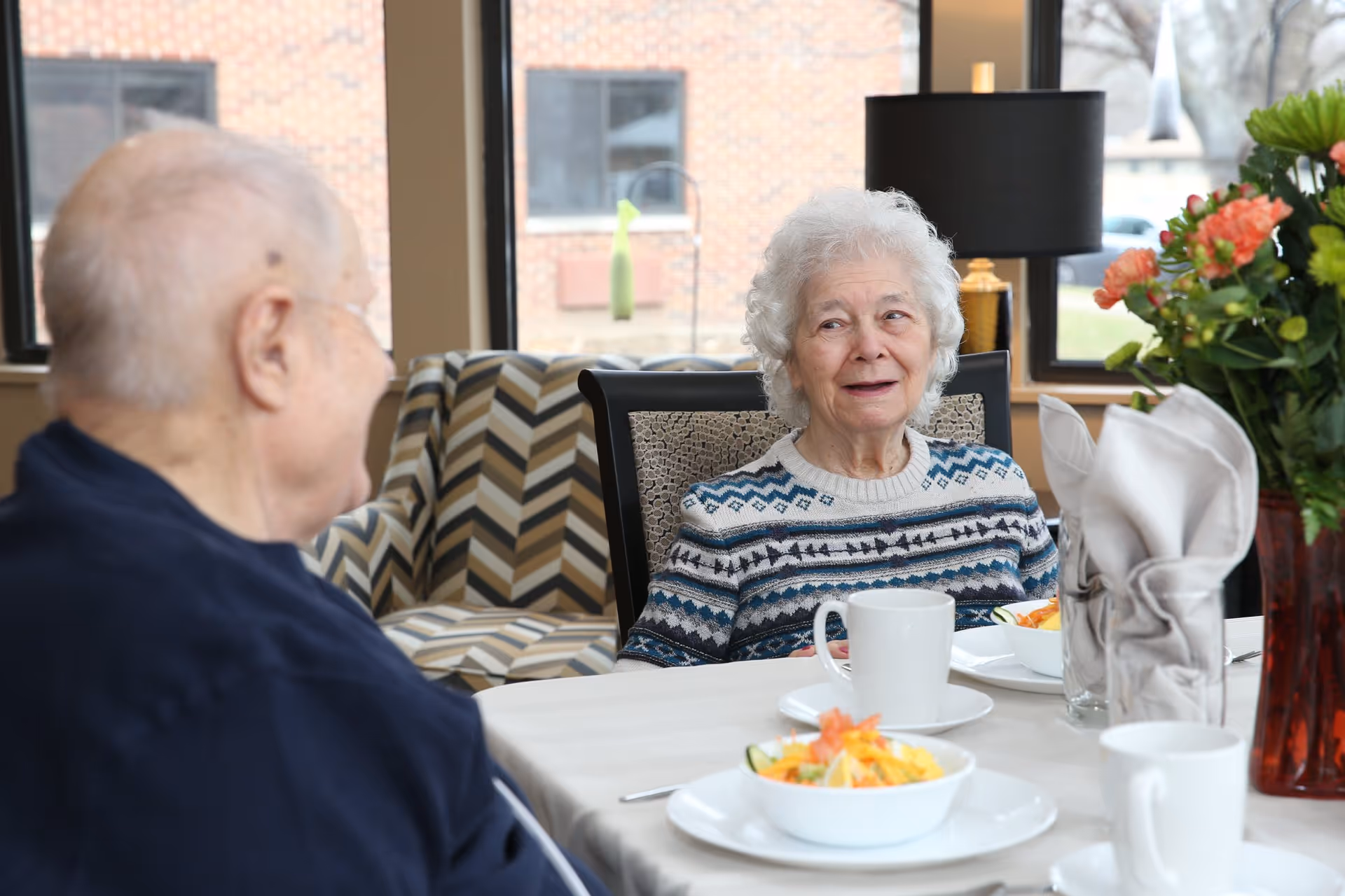 Two elderly people sitting at a dining table in a well-lit room with large windows. The woman with white curly hair is wearing a patterned sweater and looking at the man, who is seen from behind. The table is set with white cups, plates, bowls of salad, folded napkins, and a vase with flowers.