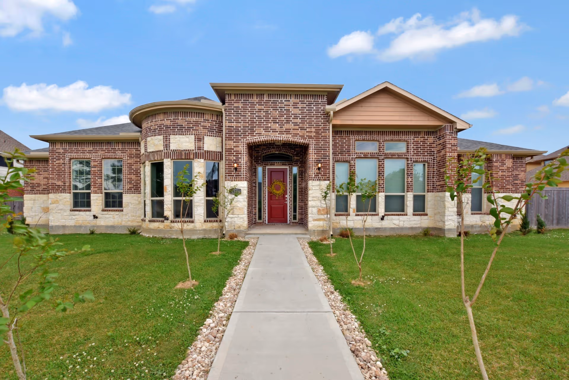 Front exterior view of a single-story brick and stone building with a red door decorated with a wreath, a concrete walkway leading to the entrance, small trees planted on either side of the walkway, and a blue sky with scattered clouds.