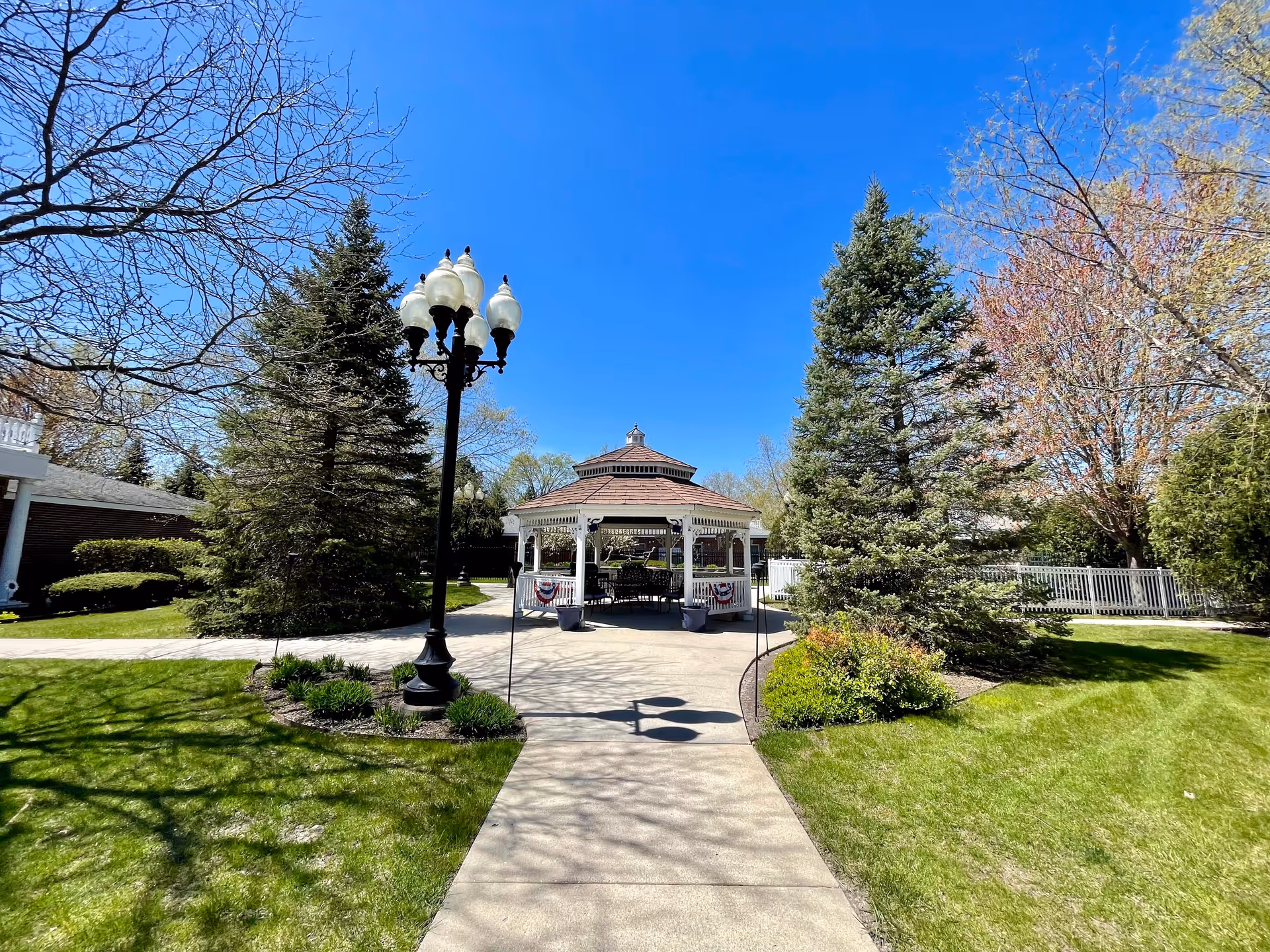 A paved walkway leads to a white gazebo decorated with patriotic bunting, surrounded by green grass, trees, and shrubs under a clear blue sky. A vintage-style street lamp stands to the left of the walkway.
