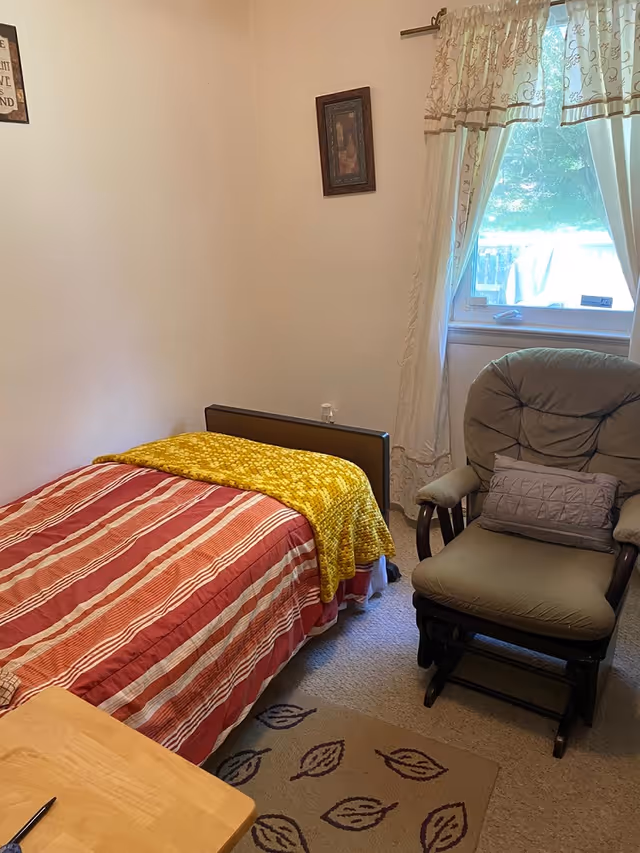 Small bedroom with a single bed covered in a red-striped comforter and yellow throw, a cushioned recliner by a curtained window, and a small table.