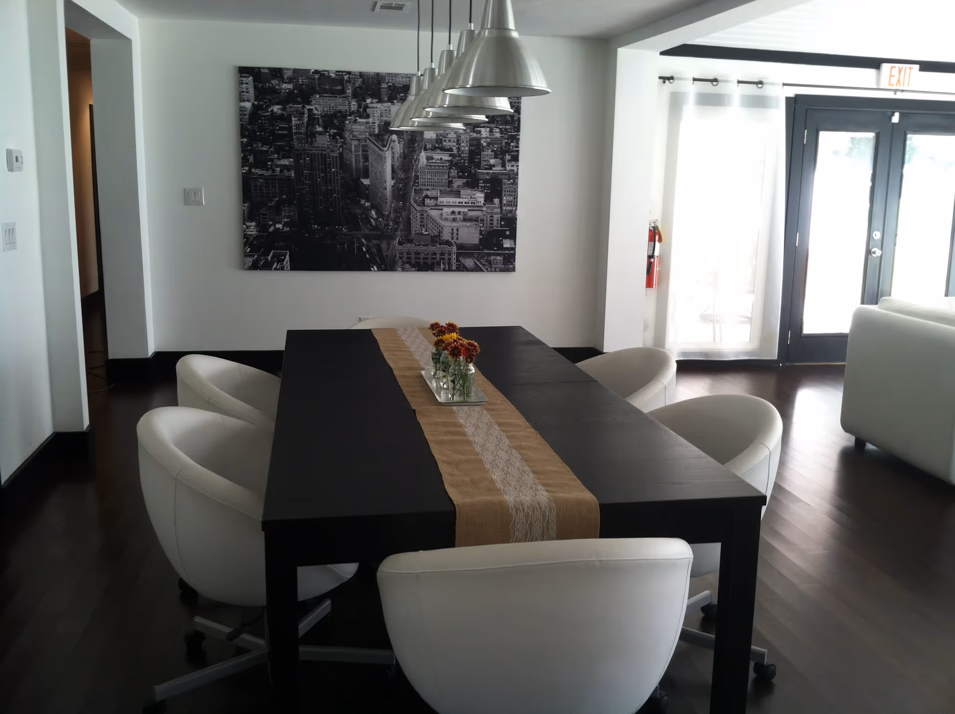 Modern dining area with a long black table covered by a beige and white lace runner, surrounded by six white cushioned swivel chairs. Above the table hang six metallic pendant lights. A black and white cityscape photograph is mounted on the white wall. The room has dark wooden flooring and large glass doors with sheer white curtains leading outside.