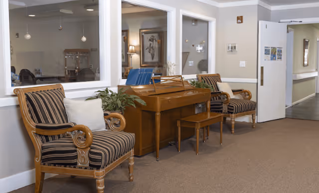 A hallway area in a senior living facility with two wooden armchairs upholstered in striped fabric, a wooden piano with a matching bench, and some green plants. The walls are light-colored with white trim, and there are large windows looking into an adjacent room. The hallway extends to the right with a door open to another area.