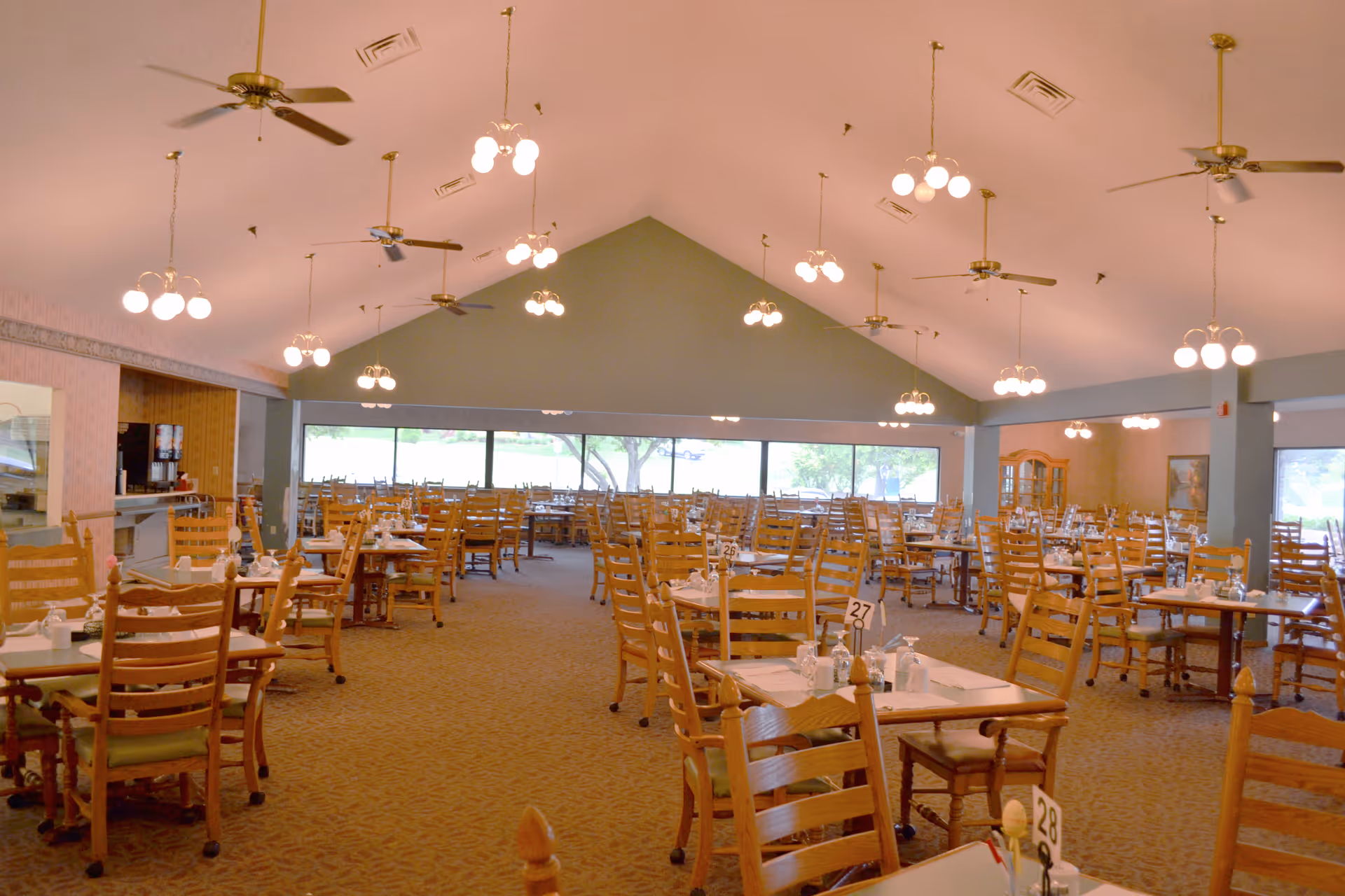 A spacious dining room with numerous wooden tables and chairs arranged neatly. Each table is set with placemats, glasses, and condiments. The room features a high vaulted ceiling with multiple ceiling fans and hanging light fixtures. Large windows along one wall provide natural light and a view of greenery outside.