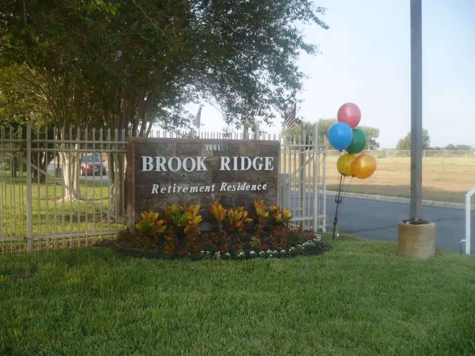 Entrance sign for Brook Ridge Retirement Residence surrounded by a small garden with colorful plants, a metal fence, a tree in the background, and a cluster of colorful balloons tied to a pole on the right side.