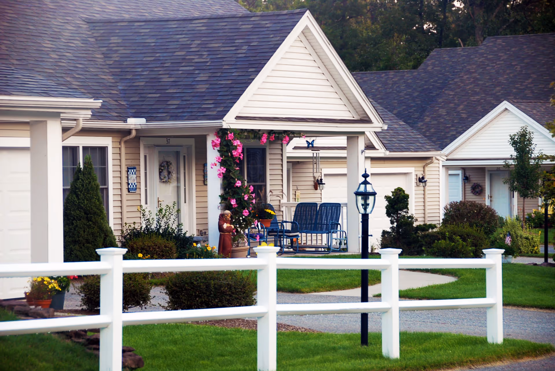Exterior view of single-story residential units with beige siding and dark shingled roofs. Each unit has a small porch with chairs, decorative plants, and flowers. A white fence runs along the front, and there is a black lamp post near the walkway. The area is landscaped with green grass, bushes, and small trees.