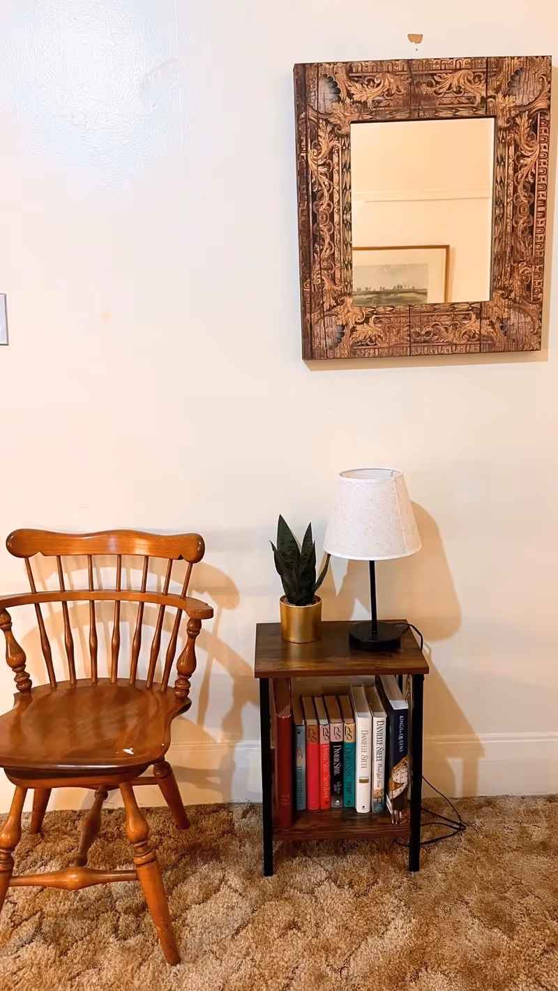 A cozy corner with a wooden chair next to a small wooden side table. The table holds a small potted plant and a lamp with a white shade. Below the table are several books neatly arranged. Above the table hangs a decorative wooden-framed mirror on a plain light-colored wall. The floor is covered with a brown textured carpet.