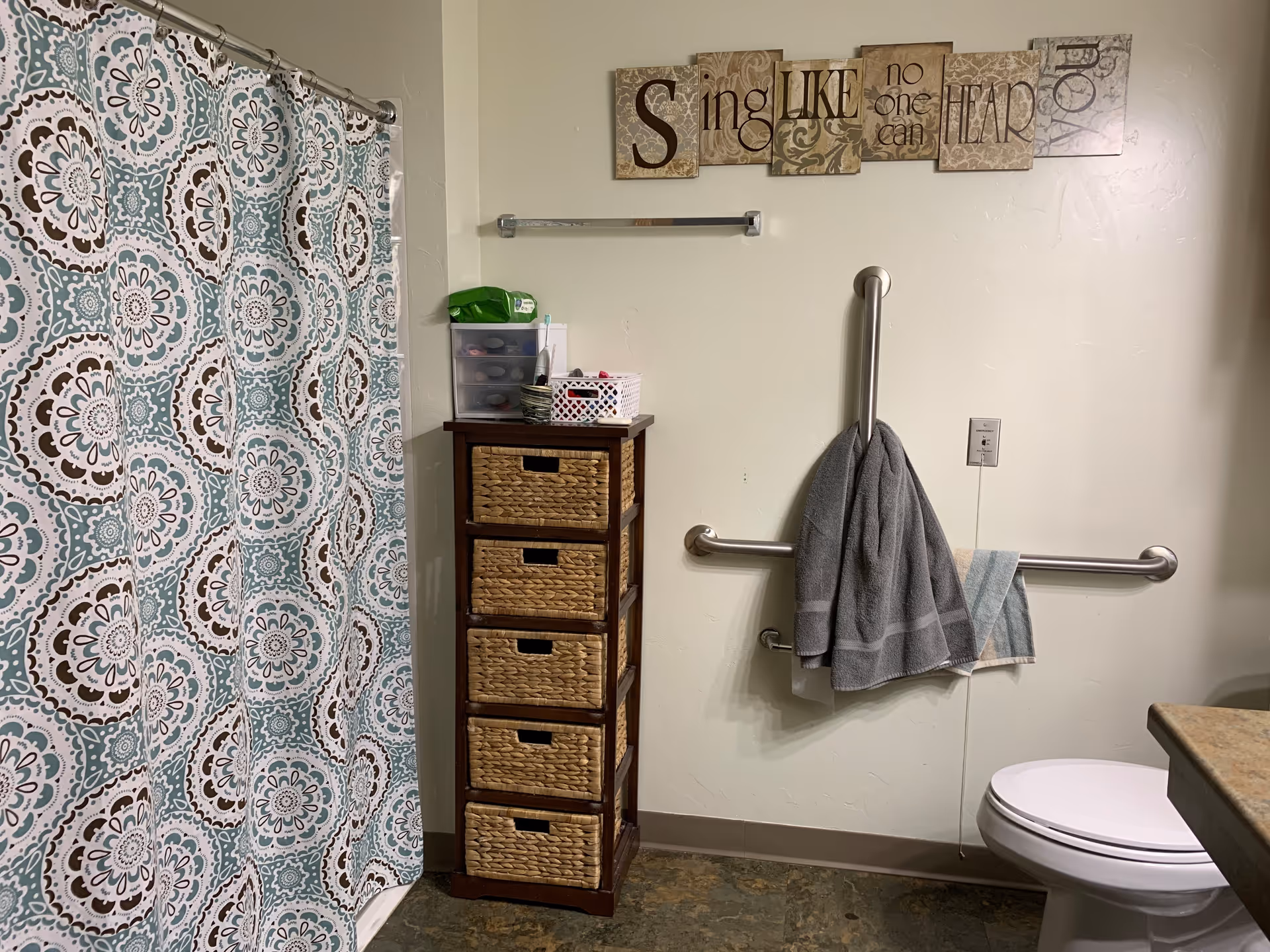 Bathroom with a patterned shower curtain on the left, a tall wooden storage unit with woven drawers next to the shower, and a toilet on the right. There are two grab bars on the wall, one vertical and one horizontal, with two towels hanging on them. Above the grab bars is a decorative wall art with the phrase 'Sing like no one can hear you'.
