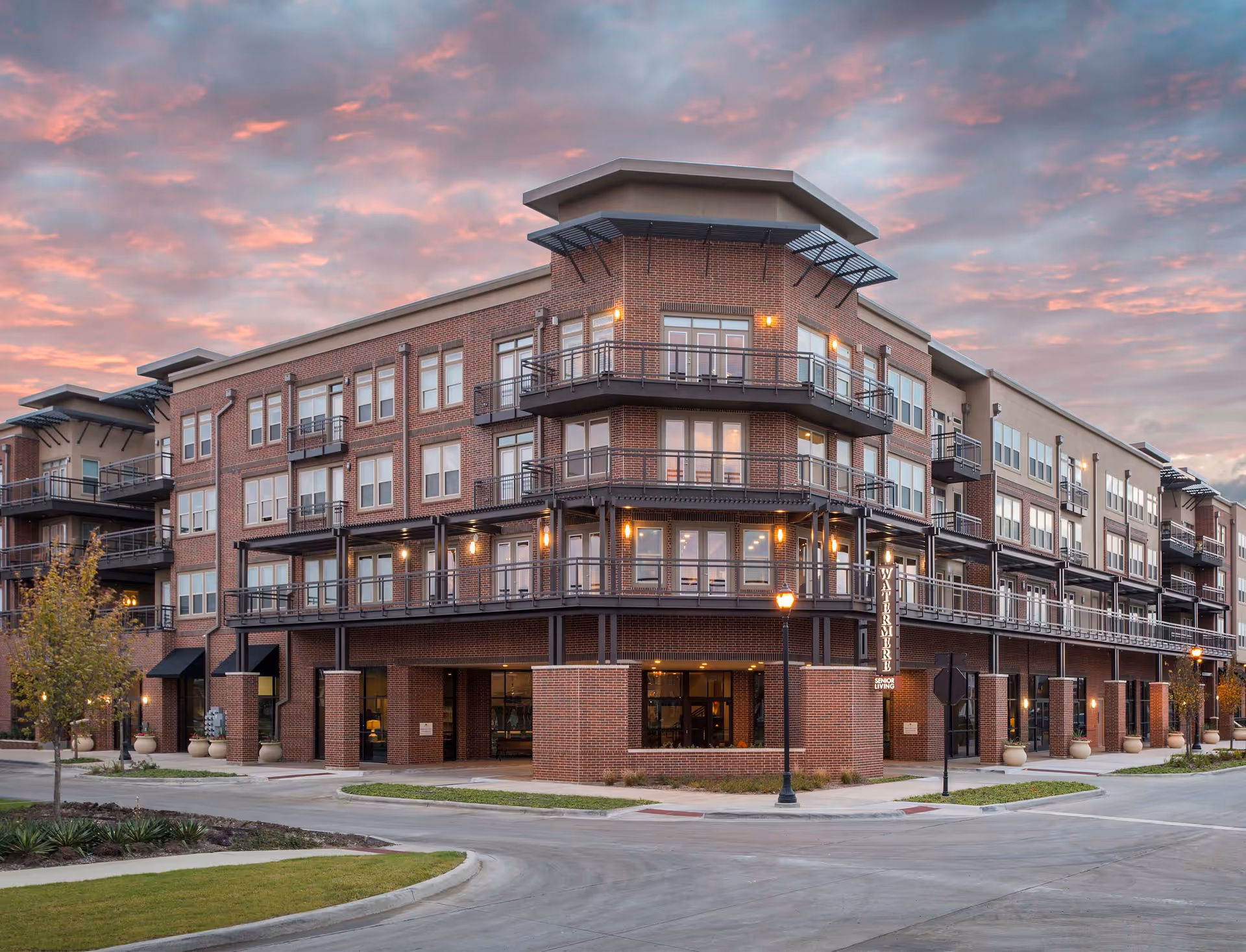 Exterior view of a multi-story brick building at sunset with balconies and large windows. The building has a sign that reads 'Watermere Senior Living' and is surrounded by a paved road and landscaped greenery.