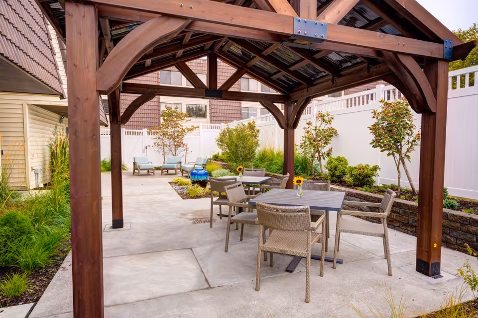 Outdoor patio area with a wooden pergola covering a table and chairs. The patio is surrounded by plants, small trees, and a white fence. Additional seating is visible in the background near a blue decorative water feature.