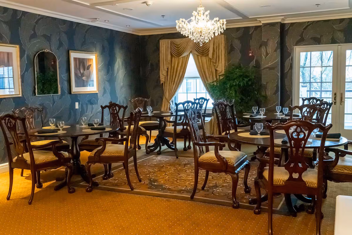 Elegant dining room with dark floral wallpaper, multiple round wooden tables set with placemats and glassware, ornate wooden chairs with cushioned seats, a crystal chandelier hanging from the ceiling, framed artwork and a mirror on the wall, large window with gold drapes, and a potted plant near glass doors.