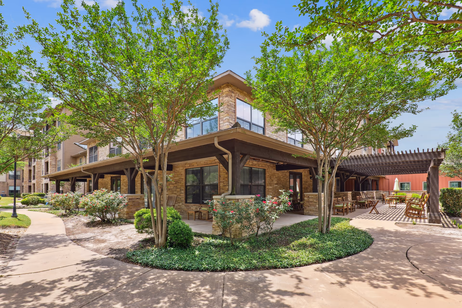 Courtyard exterior of a stone-clad senior living building with a covered patio, pergola seating, trees, and paved walkways.